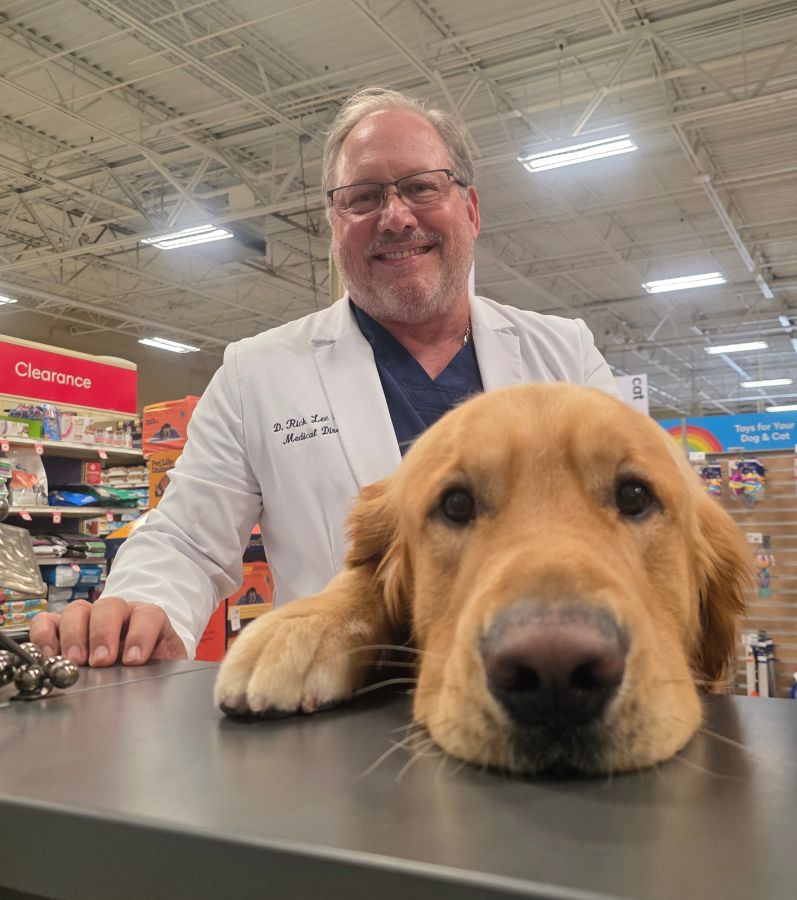 Smiling man in lab coat with a friendly golden retriever at a store counter.